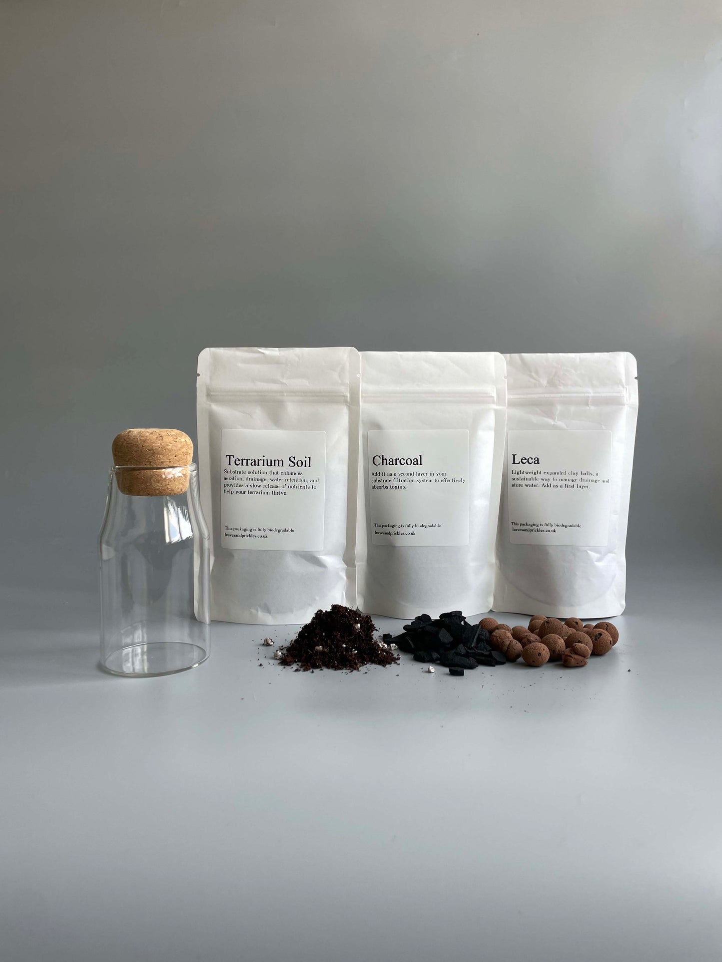 Small glass bottle with cork lid and a set of white bags labelled 'Terrarium Soil', 'Charcoal', and 'Leca on a grey background.