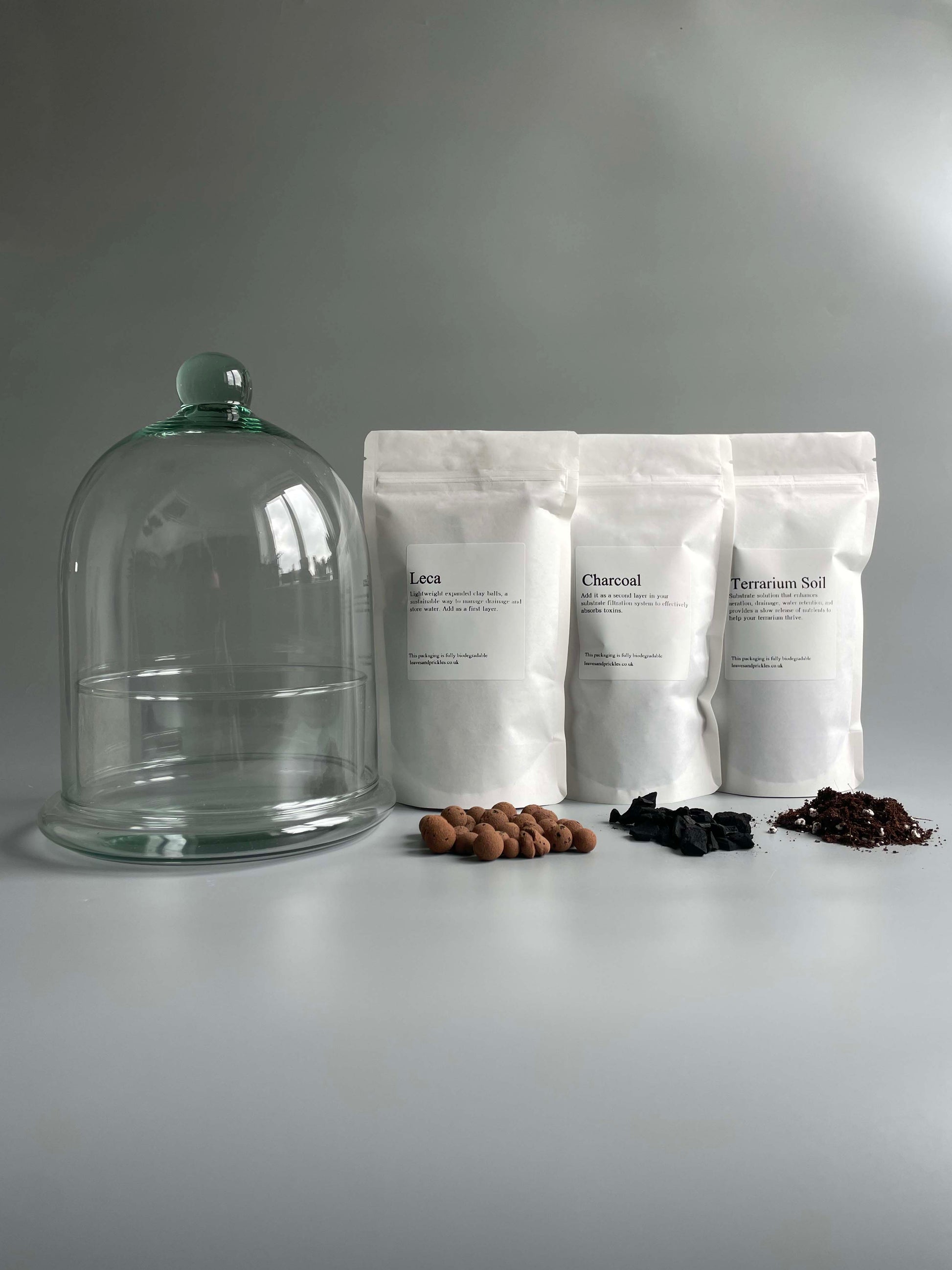 Three white bags labelled 'Leca', 'Charcoal', and 'terrarium Soil' with a glass dome on a grey background.